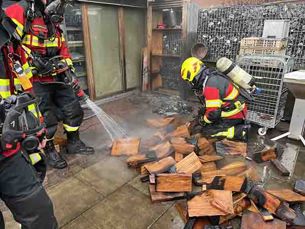 Feuerwehrleute spritzen Wasser auf Holzscheiterbeige auf einer Terrasse.
